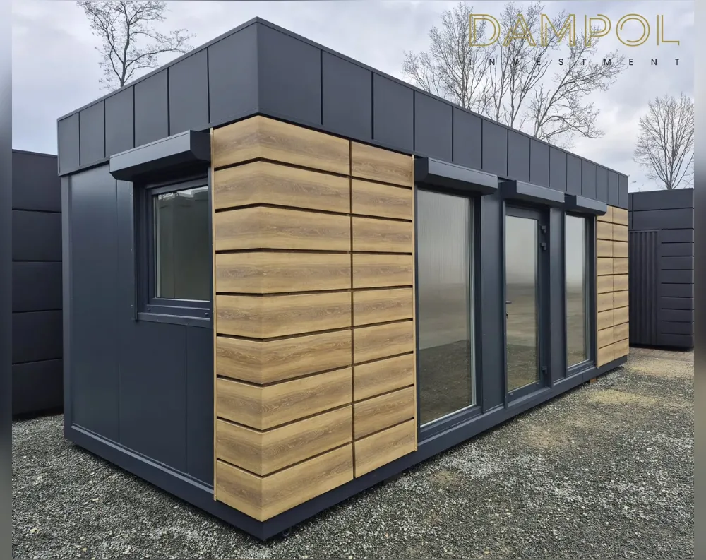 This modular building is a sleek, elongated rectangular structure with a low-slanted, mono-pitch roof that angles gently from front to back, finished in dark charcoal-grey paneling. The same graphite tone covers most of the exterior walls, contrasted by warm, horizontal wood-textured cladding panels positioned at the corners and between the windows, adding a natural accent. The front-facing side features a large glass door flanked by expansive floor-to-ceiling windows, while the side wall has one square window near the front corner. Slim dark window and door frames match the roofline trim. The structure sits on gravel ground, with bare trees and additional grey modular units visible in the background.