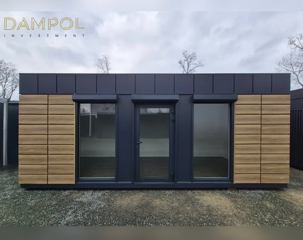 This modular building is a long, low rectangular structure with a flat roof, giving it a sleek, contemporary profile. The upper band of the façade is finished in dark charcoal-grey paneling, while the lower section combines the same dark tone with warm, horizontally slatted wood cladding on both the far left and far right sections. Centered on the front face are two large fixed glass windows flanking a dark-framed glass entry door with a black handle, all topped by a slim overhanging awning strip. The overall proportions are wide and single-story, with a low black plinth base running along the bottom, giving the structure a grounded, modern cabin-like appearance.