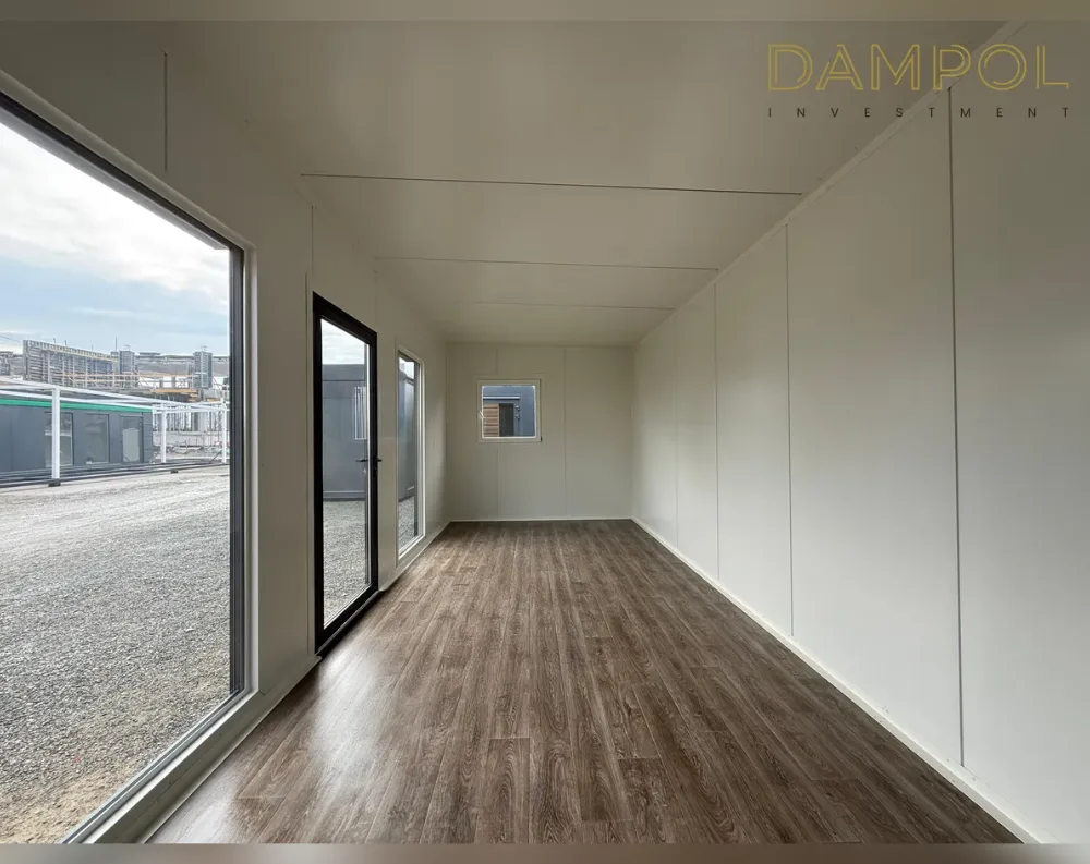 This is an interior view of a long, narrow modular room with a corridor-like proportion, stretching from the foreground toward a far wall. The walls and ceiling are finished in smooth off-white panels with subtle seam lines, giving a clean, minimalist look. The floor is covered in dark walnut-toned wood-effect flooring with a herringbone-style plank pattern. On the left side, a large fixed floor-to-ceiling window offers an expansive view of a gravel lot and distant buildings, paired with a black-framed glass entry door beside it. Further along, a second glass door and a small square window are set into the side wall, adding extra light. The far end wall is solid and windowless, closing off the space simply.