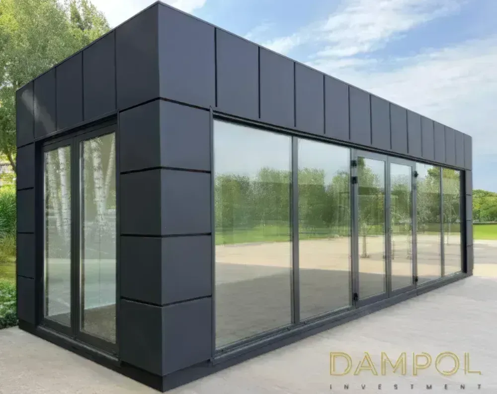 This modern modular building presents a sleek rectangular form with a low-profile flat roof and dark charcoal-grey paneled cladding covering the upper facade and corner sections in vertical segmented strips. The structure is elongated, viewed from an angle that reveals both its shorter end and long side. On the narrow end, a pair of glazed double doors sits within a dark frame. The extended side features a continuous run of floor-to-ceiling glass panels, including sliding and hinged doors with slim black frames, creating an almost fully transparent wall. The building rests on a light concrete patio, surrounded by trees and greenery, with reflections of foliage visible across the glass.
