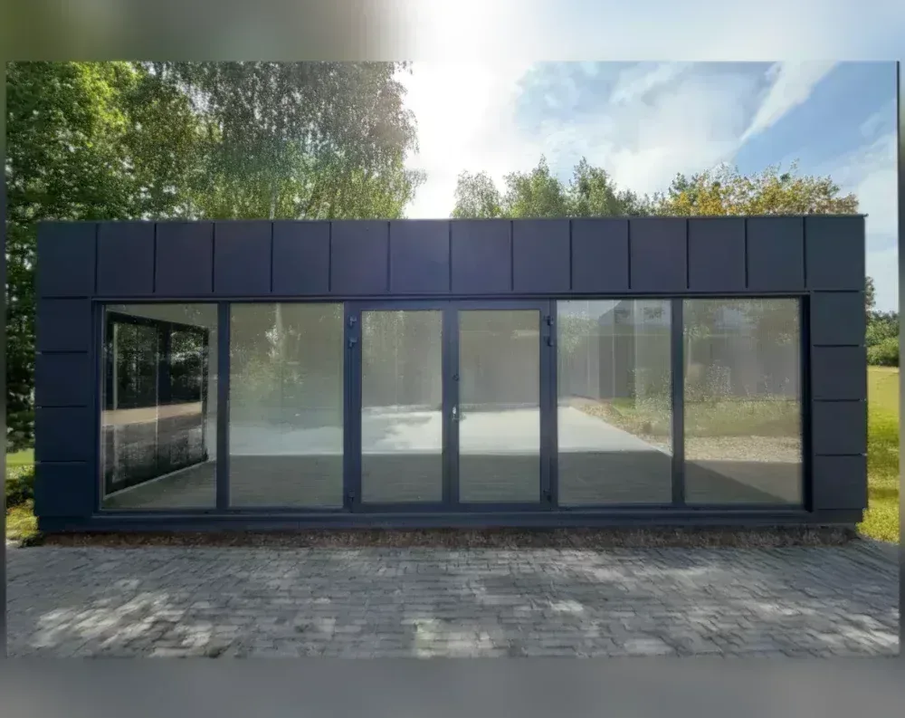 This modern garden office is a sleek, single-story rectangular structure with a low, flat roofline, appearing wider than it is tall. The exterior is clad in dark charcoal or black vertical paneling, giving it a crisp, contemporary look. The front façade is dominated by expansive glass panels: a fixed window on the far left, a large sliding glass section, a central double door with black frames, another sliding panel, and a final fixed window on the right—six glass divisions in total. The building sits on a raised base atop a stone paver patio, surrounded by lush green trees and grass, with soft sunlight illuminating the scene from above.