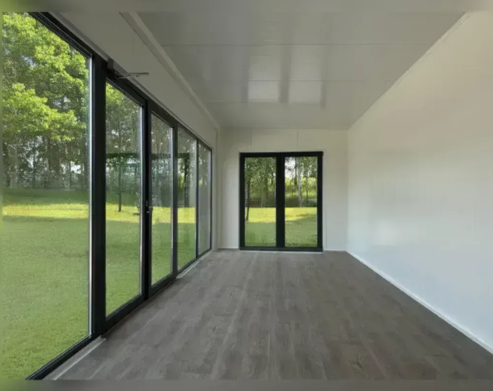 This is an interior view of a bright, empty garden office or modular building, showcasing its spacious single-room layout. The left wall features a full-length run of black-framed glass panels—appearing to be four large sliding or fixed sections—offering an expansive view of a green lawn and trees outside. At the far end, a black-framed double door with glass panels provides an additional exit point, also facing the grassy landscape. The interior has smooth white walls and a matching white paneled ceiling, creating a clean, minimalist look. The flooring is dark grey wood-effect laminate, running the length of the room, adding warmth and contrast to the bright white interior finishes.