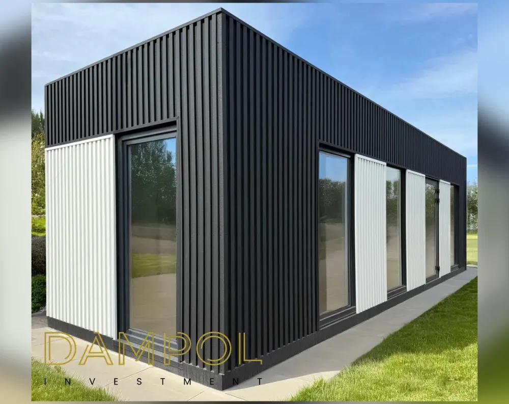 This modern modular building has a long, rectangular footprint with a sleek mono-pitch roof that slopes gently from the tall front corner down toward the rear. The exterior is clad entirely in dark charcoal vertical slat panelling, offset by contrasting white vertical slats framing the window sections. On the front-facing short side, a full-height glass door sits within a white-paneled recess. Along the long side, three large windows are evenly spaced, separated by white slatted panels, with a glazed door positioned toward the far end. The structure sits on a light concrete base, surrounded by trimmed green lawn, with trees visible in the background under a clear blue sky.