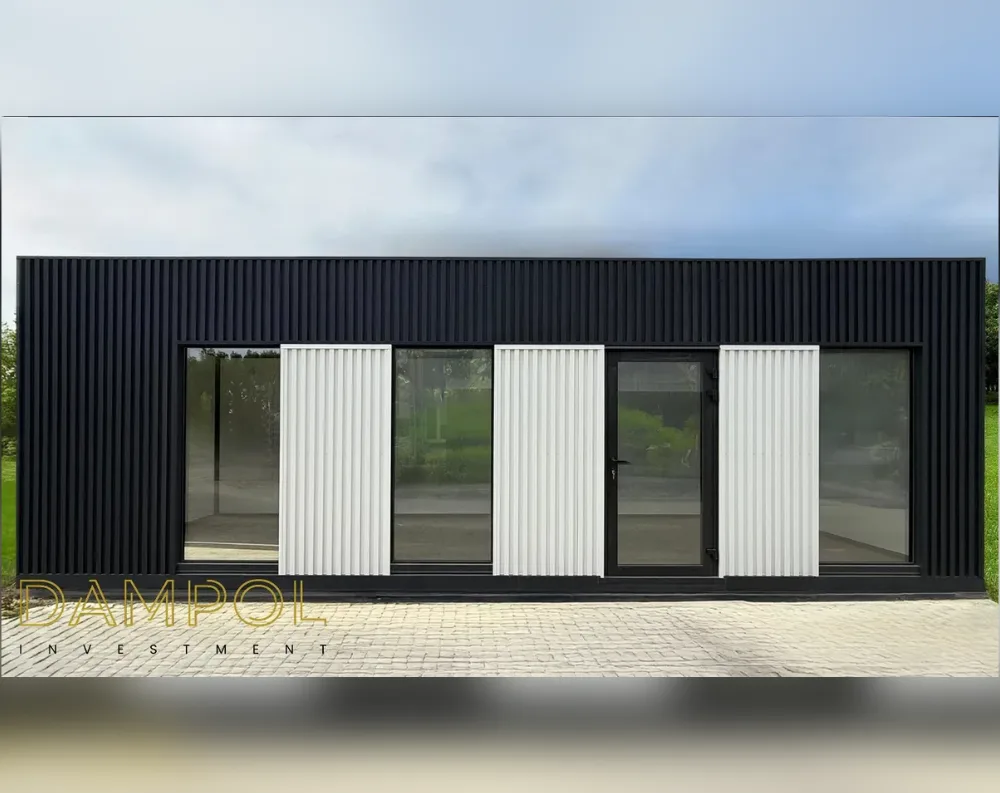 This modern modular building presents a long, low, rectangular silhouette with a flat roofline, clad entirely in dark charcoal-black vertical slat panelling that runs the full height of the structure, giving it a sleek, ribbed texture. The front façade features a symmetrical arrangement of large glass panels: two expansive fixed windows anchor each end, with a central glazed door flanked by additional glass sections. Interspersed between these are three white vertical louvre shutters, adding contrast and softening the dark exterior. The building sits atop a paved stone patio, with greenery and trees visible in the background, and a cloudy sky above completes the scene.