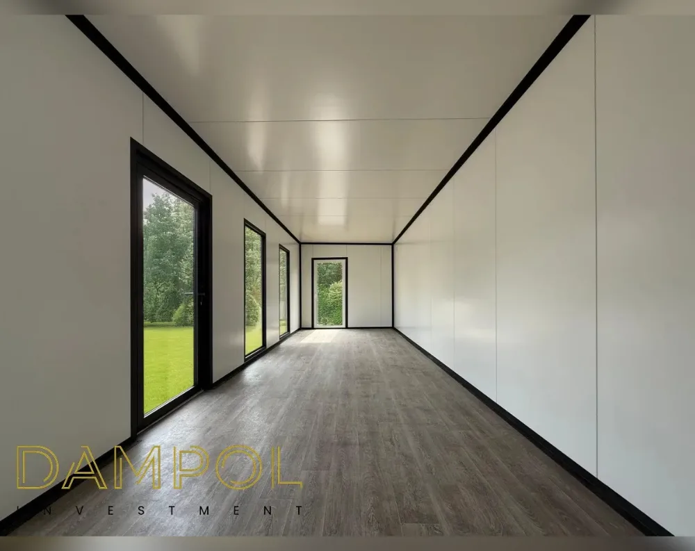 This is the interior of a long, narrow modular room with a clean, minimalist design. The space stretches back to a distant end wall, creating strong perspective lines along the dark wood-toned floor and white paneled ceiling, both trimmed with black edging that outlines every seam. On the left side, three tall glass doors and windows with black frames let in natural light and reveal a green lawn and trees outside. At the far end, a single black-framed glass door serves as the exit, flanked by white wall panels. The right-hand wall is a solid, unbroken expanse of white paneling, emphasizing the room's clean, elongated symmetry and simple, contemporary style.