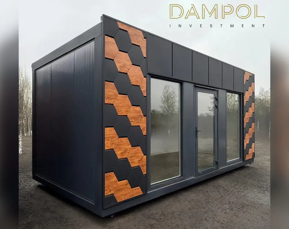 This compact modular cabin has a sleek rectangular shape with a flat roof, sitting low to the ground on small caster wheels. Its exterior is clad in dark charcoal-black paneling with a matte, textured finish. A diagonal pattern of hexagonal wood-toned tiles cuts across both the front-left and side corners, adding warmth and visual interest against the dark backdrop. The front facade features a wide glass panel arrangement: two large fixed windows flank a central glazed door with a slim vertical handle, all set within dark frames. The structure is elevated slightly on a raised platform base, and the roofline remains clean and unadorned, reinforcing the building's minimalist, modern aesthetic.