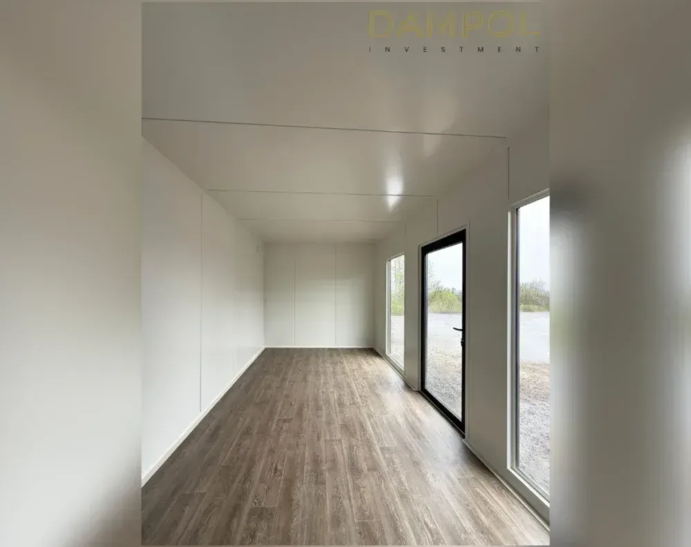 This is an interior view of a modular garden office, looking down its length toward the far end. The space is long and narrow, with a flat white ceiling and matching white wall paneling throughout, giving it a clean, minimalist feel. Recessed lighting is set into the ceiling. The flooring is a warm, wood-grain laminate in a medium brown tone, running the full length of the room. On the right-hand wall are two large fixed windows flanking a dark-framed glass entry door with a black handle, all offering views of a gravel yard and greenery outside. The left wall appears solid, without visible windows, emphasizing the room's depth and symmetry.