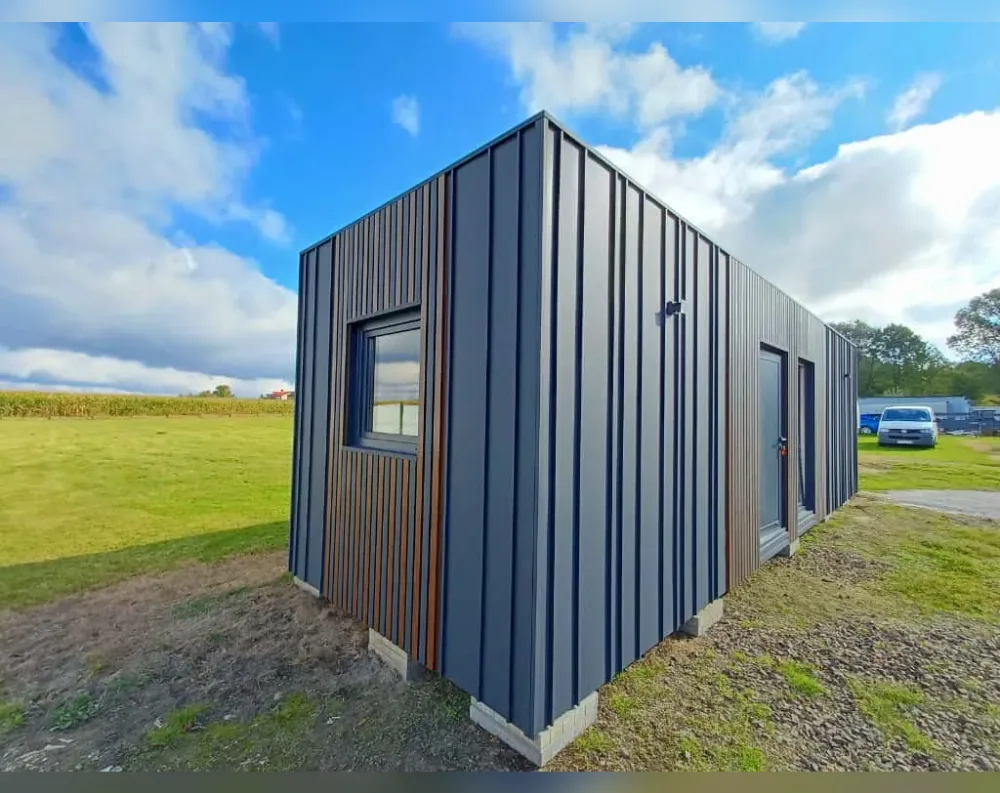 This modular garden building stands as a sleek rectangular structure with a low-pitched, single-slope roof that gives it a modern, minimalist silhouette. Its exterior is clad in vertical dark charcoal-grey metal paneling, creating clean linear texture across every surface. A single square window with slim black framing sits near the left corner, accented by a strip of warm wood-toned paneling running vertically beside it. On the opposite end, a tall glass-paneled entry door pairs with a narrow sidelight window, both trimmed in matching dark framing. A small dark light fixture is mounted on the wall between the window and door. The building rests on a low concrete block foundation, set against an open grassy field under a bright blue sky.