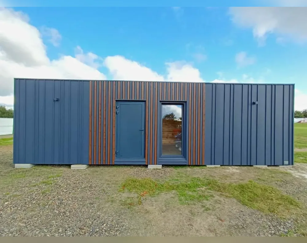 This modern modular building is a long, low rectangular structure with a flat roofline, resting on small concrete support blocks along a gravel base. Its exterior is clad mostly in dark slate-blue vertical metal panelling, with a central section featuring warm wood-toned vertical slats that create a striking contrast. Within this wood-accented middle portion sits a single solid door, painted matching blue-grey, alongside a tall narrow window with a slim frame, positioned side by side. The rest of the façade on either side is unbroken paneling, save for two small dark handles or vents high on the walls. The overall look is sleek, minimalist, and contemporary, set against a bright sky with scattered clouds.