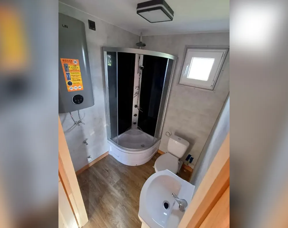 This photo shows the interior of a compact bathroom rather than the building's exterior. The small rectangular room features a corner shower enclosure with curved glass and black side panels, positioned beneath a square ceiling light fixture. A wall-mounted water heater sits to the left, above exposed pipes. A single small square window with a white frame is set into the right wall, allowing natural light in. The floor is laid with wood-effect vinyl planks. A white toilet and a corner-style pedestal sink with a chrome tap are positioned along the right side, near a wooden door frame leading out of the space.