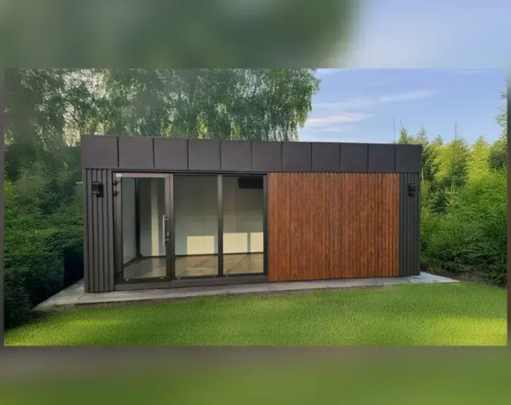 This modern garden office is a low, wide rectangular structure with a flat roof, sitting on a paved patio surrounded by lush green lawn and trees. The left two-thirds features a striking facade combining vertical black slatted panels with a large glazed opening—one hinged door and two sliding glass panels—framed in dark bronze/black metal, allowing a full view into the empty interior. The right third is clad in warm reddish-brown wood slats, running vertically, contrasting with the black paneling above and around it. The upper band above the glass and wood sections is finished in smooth dark charcoal panels. Small black wall-mounted lights flank the entrance area.