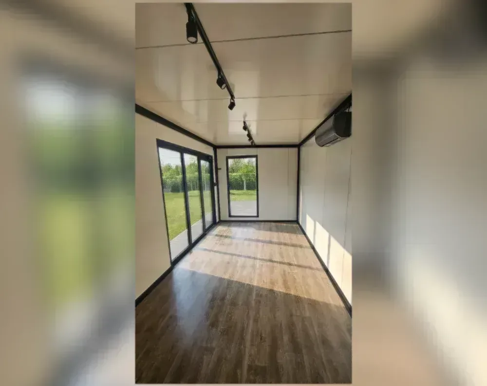 This is an interior view of a compact modular garden office with a rectangular footprint and a flat ceiling. To the left, a large sliding glass door system with three panels in black aluminum framing opens onto a grassy lawn. Adjacent to it, on the back wall, sits a tall rectangular window, also black-framed, looking out to a wooden deck and hedged greenery beyond. The walls are finished in smooth cream-colored paneling with dark trim seams marking the panel joints and corners. The floor is light wood-toned laminate with subtle gray accent strips running across it. A modern split-unit air conditioner is mounted high on the right wall, and track lighting spans the ceiling.