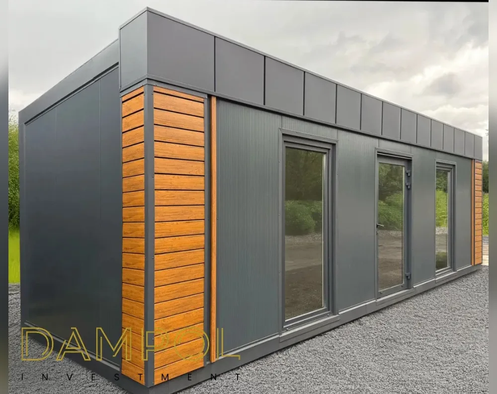 This modular building is a long, rectangular structure with a low-profile flat roof, photographed at an angle that shows both its front and side facades. The exterior is finished in dark charcoal-grey paneling with vertical seams, giving it a sleek, contemporary look. Warm honey-toned horizontal wood slat accents frame the corners and edges, breaking up the grey with natural texture. Along the front face are two large glass window panels flanking a central glass-paneled door, all set in matching dark grey frames. The structure sits on a bed of grey gravel, with greenery and an overcast sky visible in the background, reinforcing its use as a standalone garden office or cabin.