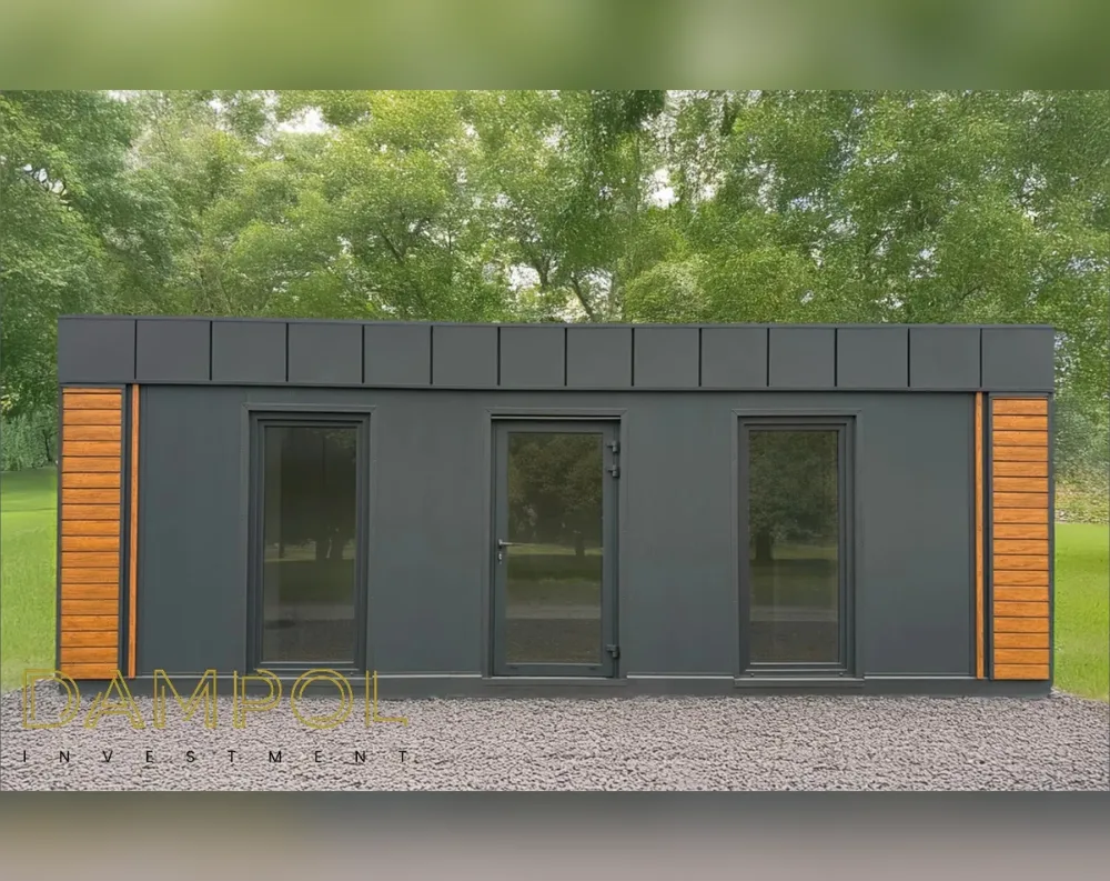 This modern modular structure has a long, rectangular, single-story footprint with a flat roof edged by a paneled fascia band running its full length. The exterior is finished in smooth dark charcoal-grey cladding, contrasted by warm horizontal wood-toned slats bookending each side of the building. The front façade features three tall, dark-framed glass openings evenly spaced: a fixed window on the left, a central hinged glass entry door with a slim handle, and another large window to the right, all matching in height and trimmed in matching dark grey frames. The building sits on a bed of light grey gravel, set against a lush green wooded backdrop.