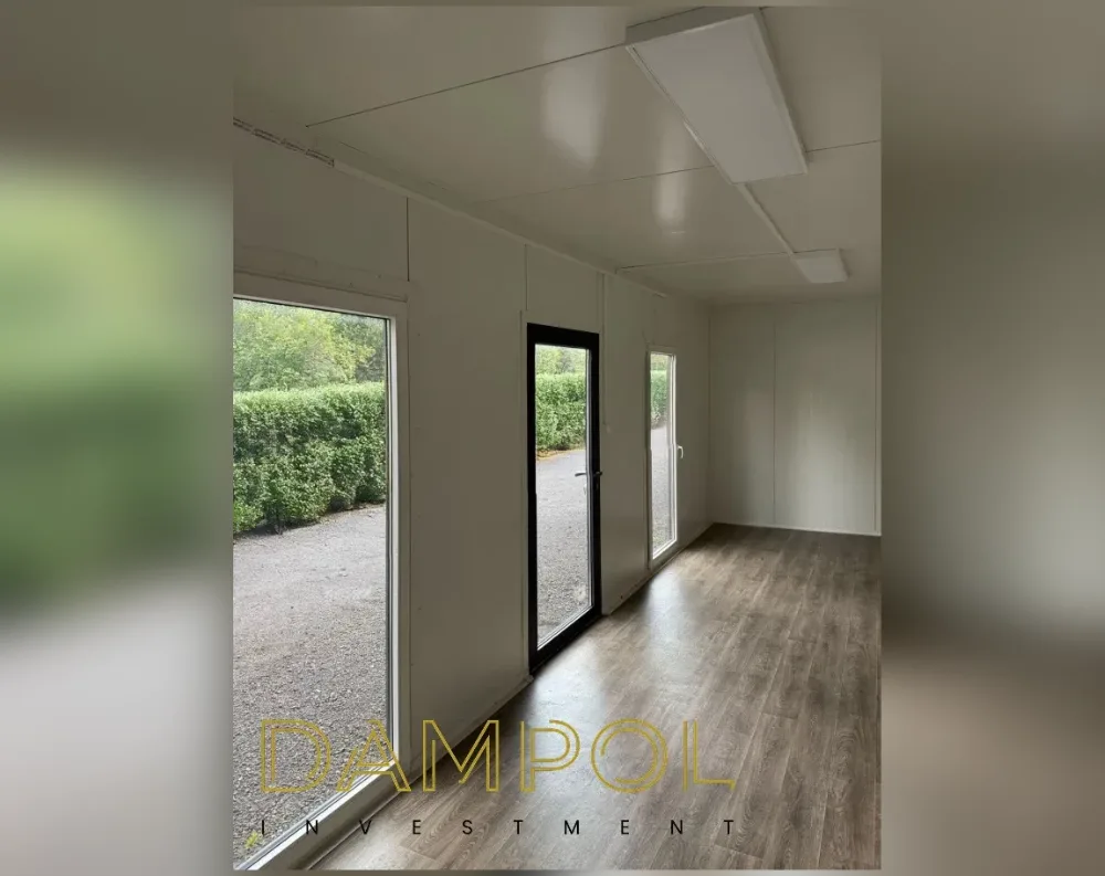 This bright, empty interior shows a narrow rectangular modular room with a long perspective from front to back. Along the left wall are two large fixed windows flanking a central glazed door with a black aluminum frame, all overlooking a gravel path and a dense green hedge outside. The walls and ceiling are finished in smooth white paneling, and an oblong LED panel light is mounted overhead, illuminating the space evenly. The flooring is warm brown wood-look laminate, running the length of the room toward a plain white far wall. The design is clean and minimalist, with the black-framed openings providing the only strong contrast against the bright white interior.