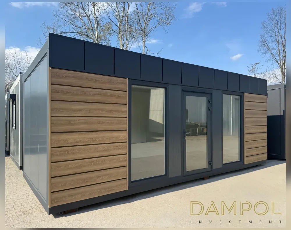 This modular building is a long, rectangular single-story structure with a flat roof and dark charcoal-grey trim running along the fascia. The front façade combines warm, wood-look horizontal slat cladding on both outer sections with a central dark-grey panel housing the entrance. That center section features a full-height glass door flanked by two large fixed windows, all framed in matching dark grey, allowing a clear view into the empty interior space. The side wall, partially visible, is finished in a lighter grey ribbed metal panel. The building sits raised slightly off the ground on a paved lot, with bare trees and a blue sky with light clouds in the background.