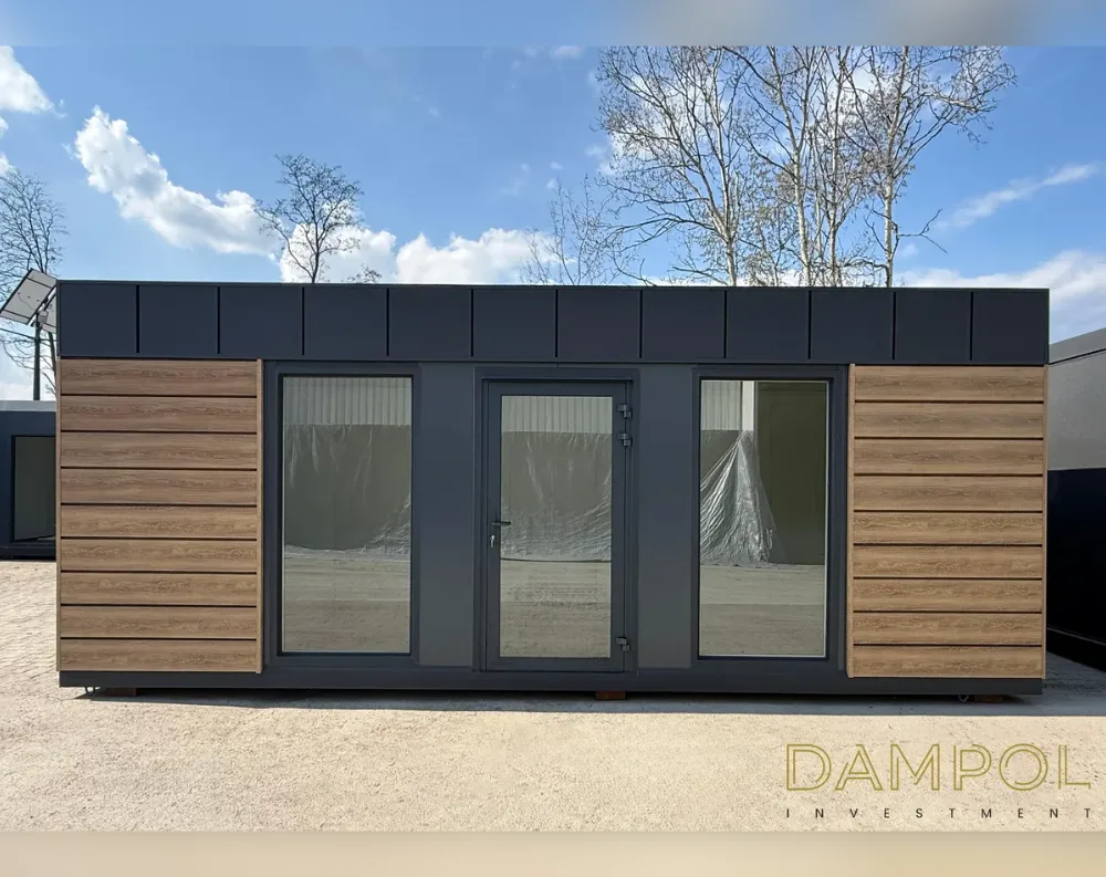 This modern modular structure is a long, low rectangular building with a flat roof, its dark charcoal fascia running the full length above the facade. The front elevation is symmetrical: horizontal wood-toned composite cladding panels bookend each side, while the central section features a glass-paneled entry door flanked by two large fixed windows, all framed in matching dark grey. The interior glimpsed through the glass appears unfinished, with protective plastic sheeting visible. The building rests on small dark support feet, slightly elevated above a gravel surface. Behind it, bare-branched trees and a solar panel edge into view against a partly cloudy blue sky, emphasizing the structure's crisp, contemporary lines and clean contrast between wood and charcoal tones.