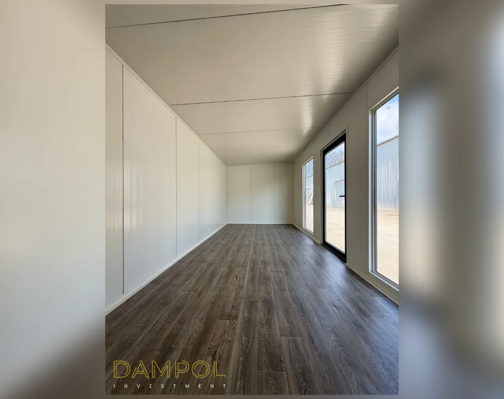 This photo shows the interior of a long, narrow modular room with a clean, minimalist finish. The walls and ceiling are clad in smooth white paneling with subtle seam lines, creating a bright, uniform look throughout. The floor is finished in dark, wood-grain laminate that runs the length of the space, adding warmth against the pale walls. At the far end, natural light floods in through a glass-paned entry door flanked by tall vertical windows on either side, offering a view of an outdoor gravel area and a neighboring structure. The ceiling is flat and low-profile, following the rectangular footprint of the building, giving the space a streamlined, functional, contemporary feel.