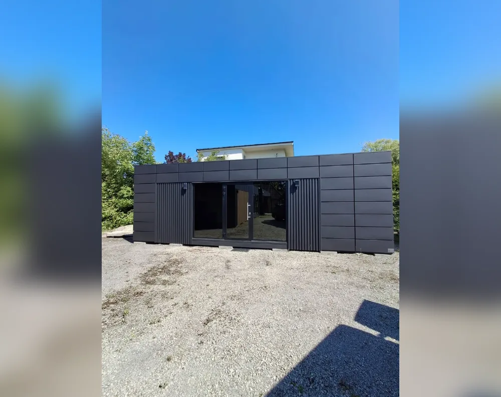 This modular garden office is a long, low, single-story rectangular structure with a flat roof, clad entirely in dark charcoal-black panels arranged in horizontal rows. Centered on the front façade is a wide glass entrance featuring double sliding doors flanked by narrow vertical slat-style panels on both sides, creating a striking textured accent against the smooth cladding. Small wall-mounted lights sit just above each side of the doorway. The building has no additional visible windows beyond this central glazed doorway section. It rests on a raised base, slightly elevated off the gravel ground, with a bright blue sky above and greenery framing both sides, while a larger house is visible in the background.
