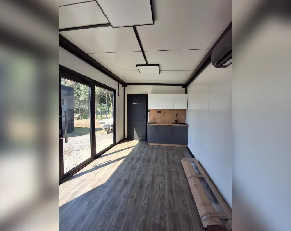 This is an interior view of a compact modular garden office or tiny cabin. The space is long and narrow, with a dark wood-effect laminate floor stretching toward large sliding glass doors on the left wall, which flood the room with natural light and reveal trees and gravel outside. A dark charcoal single door sits beside a small fitted kitchen unit with white upper cabinets, a wooden backsplash, dark lower cabinets, and a sink with black tap. The ceiling is white with recessed square panel lighting and exposed dark structural beams. A wall-mounted air conditioning unit is fitted near the ceiling. Walls are smooth white paneling, and flat-packed materials rest on the floor near the back.