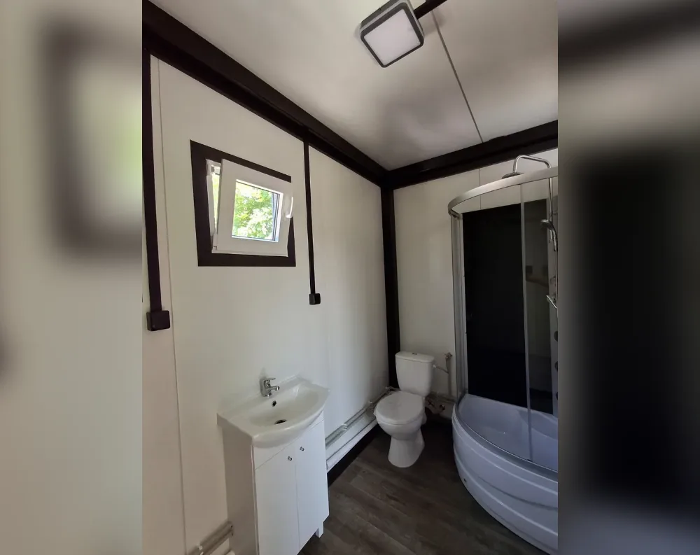 This compact modular bathroom features clean white paneled walls accented with dark brown trim strips running vertically and along the ceiling edges, giving a modern cabin-like feel. A single tilt-style window with a dark frame sits centrally on the back wall, allowing natural light and a glimpse of greenery outside. The space is efficiently laid out: a white pedestal sink with cabinet storage sits in the near corner, beside a matching white toilet. To the right, a curved glass-enclosed shower/tub combination with a chrome fixture fills the corner. The flooring is dark wood-toned laminate, contrasting the flat white ceiling, which holds a square light fixture and visible wiring conduit running along the beam.