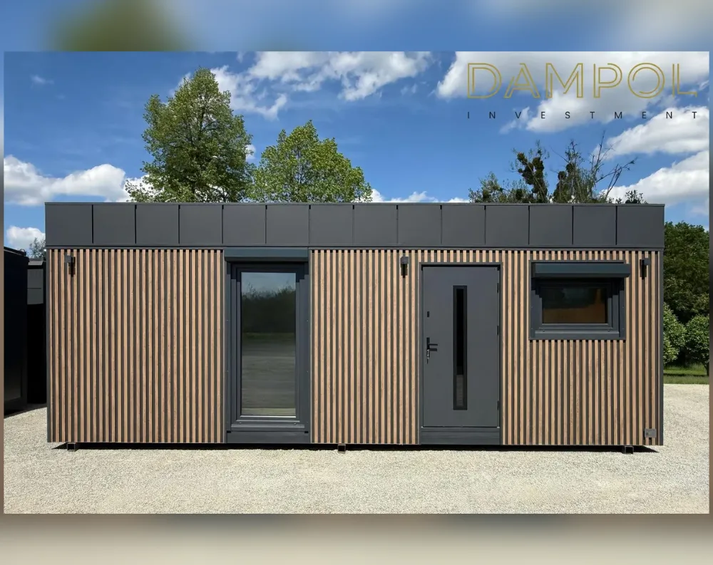 This modular garden building has a long, rectangular, single-story profile with a flat roof and dark gray fascia trim running along the top edge. The exterior is clad in vertical wood-toned slatted panels, alternating light and dark tones for a textured, contemporary look. The facade features one large floor-to-ceiling glass door on the left with slim dark framing, a centrally placed solid gray entrance door with a narrow vertical window and black handle, and a smaller rectangular window to the right with matching dark trim. Small black wall-mounted lights flank the entrance door. The structure sits on short support feet above a gravel surface, with trees and a blue sky visible behind it.