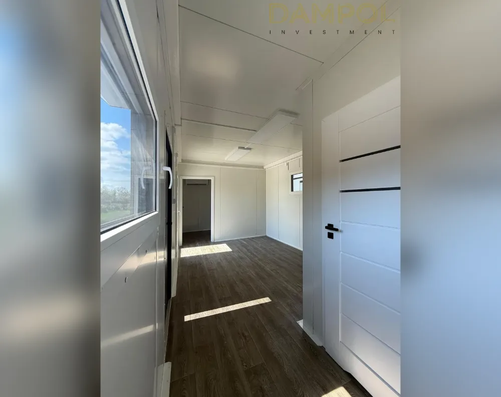 This interior shot reveals a modular room with clean, minimalist styling. Dark walnut-toned laminate flooring runs the length of the space, catching bands of natural sunlight. To the left, a large rectangular window with white frames overlooks a grassy field and blue sky, paired with a glazed door featuring a black handle. The ceiling and walls are finished in smooth white paneling, with a subtle recessed light fixture overhead. On the right, a white paneled interior door with horizontal black accent stripes and a black handle stands open. Further down the corridor, another small window and doorway lead to an adjoining room, all bathed in bright, airy light.