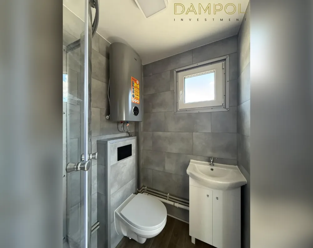 This photo shows the interior of a compact bathroom rather than the building's exterior. The space features grey stone-effect tile cladding covering the walls, with a dark wood-toned floor. A single small window with white framing sits centrally on the back wall, letting in daylight. To the left, a glass shower enclosure with a chrome handle occupies the corner, and above it a cylindrical silver water heater is mounted. A wall-hung white toilet sits beneath the heater, with visible plumbing pipes running along the wall. To the right, a white pedestal-style sink with a cabinet base stands beneath the window, completing the fixtures.
