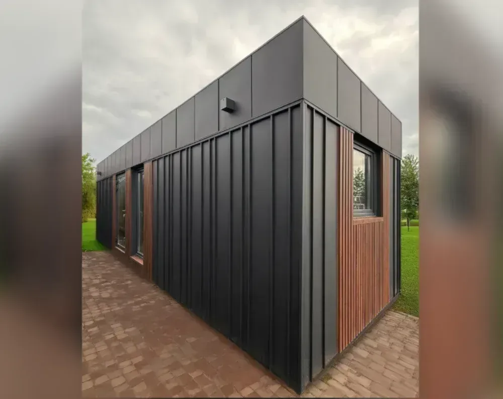 This modern garden building has a sleek, elongated rectangular form with a flat roof, capped by a black metal fascia that wraps seamlessly around the structure's edges. The exterior is clad predominantly in dark charcoal-black vertical corrugated metal panels, giving it a crisp, contemporary industrial look. Contrasting warm cedar-toned wood slats frame the entry areas, adding natural texture against the black. Along the long side, two tall windows sit beside a wood-trimmed entrance, while the near end features another large window framed similarly in timber. A small black wall-mounted light fixture sits beneath the roofline. The building rests on a herringbone brick paved patio, surrounded by green lawn and trees under an overcast sky.