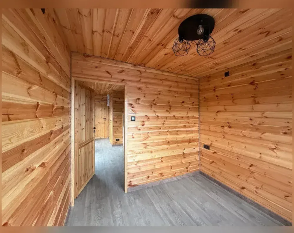 This interior view shows a cozy, wood-lined room within what appears to be a modular garden building, entirely clad in warm honey-toned tongue-and-groove pine paneling that covers the walls and ceiling alike, creating a cabin-like atmosphere. The space is rectangular, with a doorway on the left wall leading into an adjoining hallway or room, where another wooden door is visible further back. A modern black ceiling light fixture with geometric wire-cage shades hangs prominently, contrasting against the natural wood tones. Small electrical outlets and switches are mounted on the walls. The flooring is a grey-toned laminate or vinyl plank, offering a cool contrast to the warm wood surfaces surrounding it.