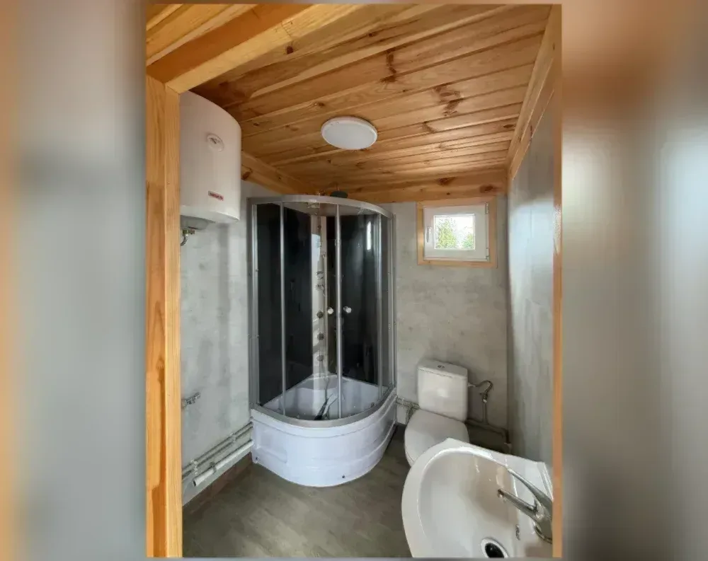 This compact bathroom sits beneath a warm, honey-toned wood-paneled ceiling with visible plank grooves, lit by a single round flush-mount fixture. A wood-trimmed doorway frames the entry, its natural timber beams contrasting against smooth grey stone-textured walls. In the corner, a curved glass-and-chrome shower enclosure dominates the space, paired with a white water heater tank mounted above on the wall, alongside visible plumbing pipes running along the baseboards. A small window with wooden trim sits high on the right wall, letting in daylight and a glimpse of greenery outside. Below it, a white toilet and the edge of a sink with chrome fixtures complete the fittings, all set atop dark wood-look flooring.