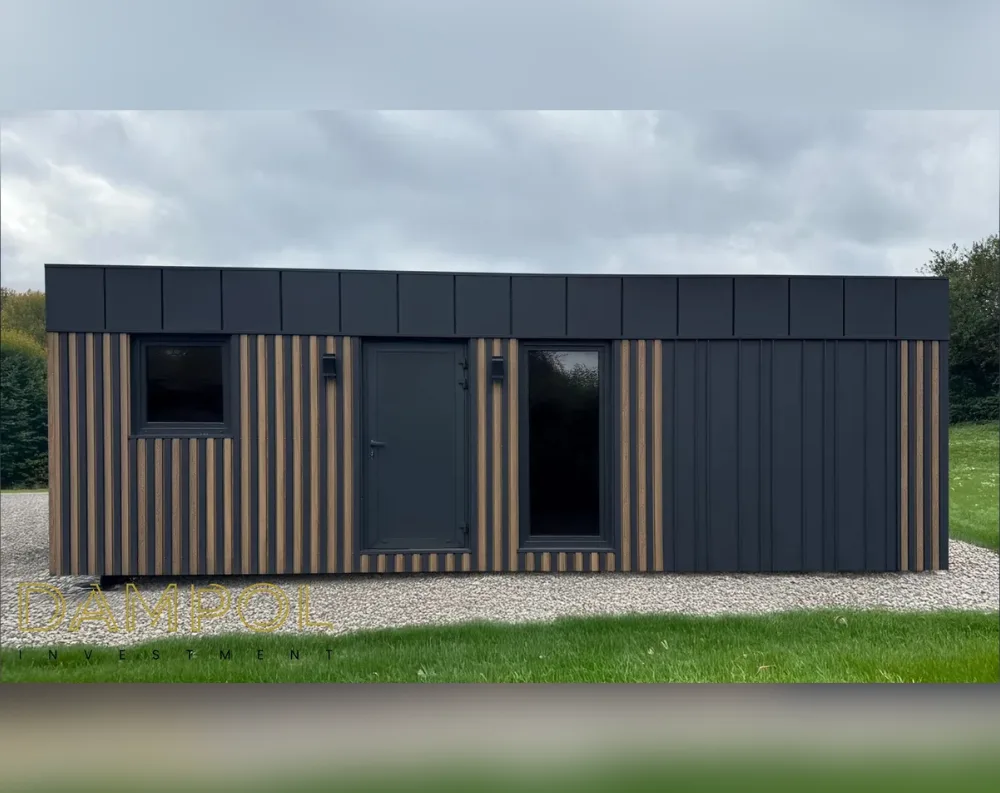 This modern garden building is a long, low rectangular structure with a flat, dark roof that has a slightly angled fascia along the top edge. The exterior combines two textures: vertical dark anthracite metal cladding on the right two-thirds, and alternating black-and-wood-toned vertical slats on the left portion. A single square window sits near the far left, followed by a dark entrance door with a small canopy light above it, and then a tall glazed window or second door beside it, both trimmed in matching black frames. The building rests on a pale gravel base surrounded by green lawn, with trees forming a backdrop under a cloudy sky.