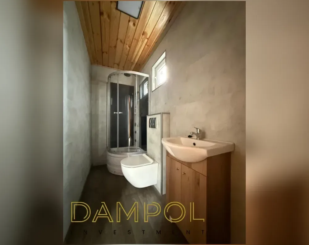 This photo shows the interior of a compact bathroom within a modular building, rather than its exterior. The narrow, elongated room features a wood-paneled ceiling with visible plank grain, transitioning to textured concrete-toned walls. A corner shower cubicle with curved glass paneling sits at the far end, illuminated by a small rectangular window mounted high on the side wall. Beside it, a wall-mounted white toilet is positioned against a tiled control panel. To the right, a pedestal-style sink with a chrome faucet rests atop wooden cabinetry with round metal knobs. The flooring is dark wood-toned laminate, and an overhead skylight adds natural light near the ceiling's peak.
